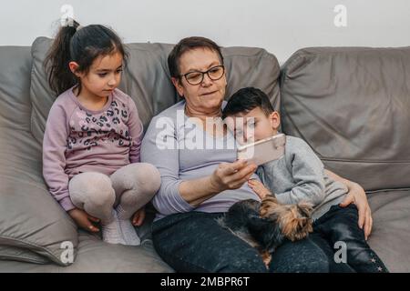 Grand-mère et petits-enfants regardant un téléphone mobile ensemble. Femme âgée, deux petits enfants et terrier du yorkshire dans un canapé à la maison. Banque D'Images