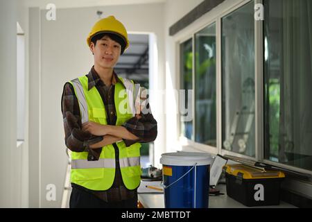 Portrait de peintre dans un casque de sécurité tenant une brosse de peinture debout sur le chantier et souriant à la caméra Banque D'Images