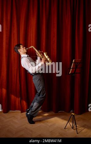 Un jeune musicien joue du saxophone ténor sur scène avec des rideaux drapés rouges Banque D'Images