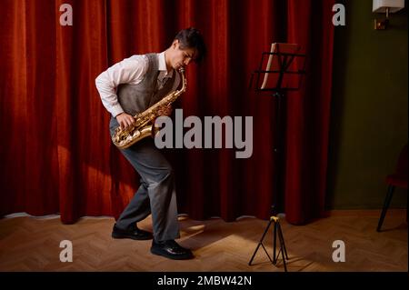 Un jeune musicien attrayant joue du saxophone ténor sur scène avec des rideaux drapés rouges Banque D'Images