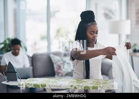 Le fait de se conformer à la routine le rend en quarantaine. une jeune femme repassant des vêtements avec son mari sur le canapé en arrière-plan. Banque D'Images