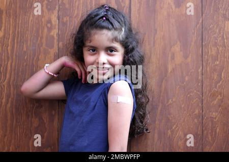 Petite fille latine aux cheveux bouclés heureux montre son bras récemment vacciné contre Covid-19 dans la nouvelle normale pour la pandémie de coronavirus Banque D'Images