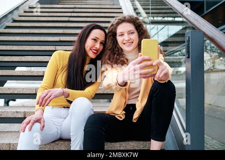 Deux femmes souriant tout en prenant des selfies avec un téléphone portable assis sur les escaliers à l'extérieur. Banque D'Images