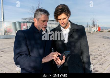 Homme d'affaires senior concentré dans des vêtements formels de navigation de téléphone mobile tout en faisant la démonstration de l'écran à ses collègues tout en se tenant debout dans la rue Banque D'Images