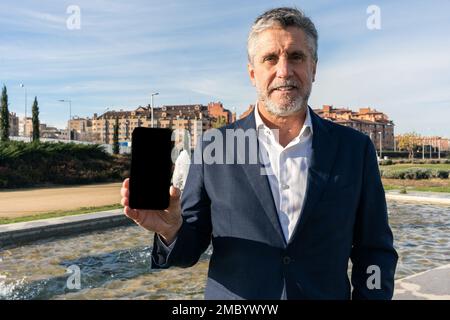 Homme d'affaires d'âge moyen positif dans une tenue habillée souriant et regardant l'appareil photo tout en montrant un smartphone avec écran noir dans la rue Banque D'Images