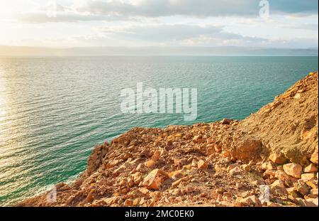 Rive de la mer morte du côté du Jourdain, plage de sable sec et rochers, le soleil brille sur la belle surface de l'eau azur Banque D'Images