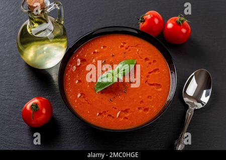 Soupe de tomate chaude et cuillère sur fond noir. Portion de purée de soupe fraîchement préparée de tomates rôties, d'ail et de basilic dans un bol noir. Nourriture végétalienne. Banque D'Images