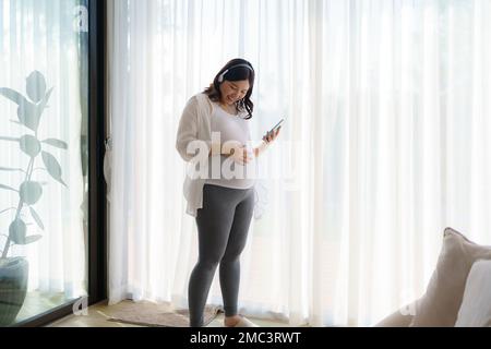 Portrait d'une femme asiatique heureuse enceinte debout dans le salon à la maison et utilisant un smartphone pour écouter de la musique dans un casque. Banque D'Images
