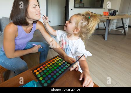 Vue latérale du jeu que la mère et la fille jouent dans le salon: Le petit met le maquillage sur elle et elle prétend être ticklich dans le nez Banque D'Images