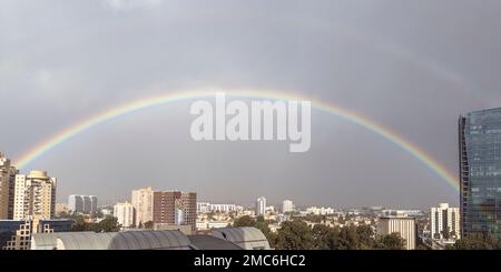 un bel arc-en-ciel plein sort d'un ciel nuageux au-dessus d'une grande ville avec quelques bâtiments élevés contre un ciel nuageux avec le soleil essayant de sortir Banque D'Images