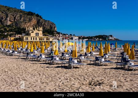 Chaises longues vides et parasols jaunes sur la plage de la petite ville de Mondello, la face rocheuse du mont Gallo derrière les maisons. Banque D'Images