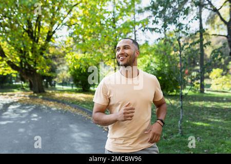 Joyeux et réussi hispanique homme jogging dans le parc, homme courant une journée ensoleillée, souriant et heureux d'avoir une activité en plein air. Banque D'Images