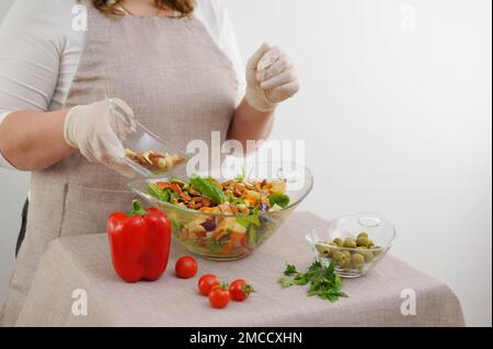 Femme en gros plan cuire en gants verse des noix dans des ingrédients de salade sur le tablier de cuisine poivre tomates persil olives verts croûtons stériles gants place pour le texte vêtements blancs Banque D'Images