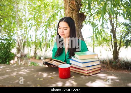 Jolie jeune femme lisant des livres sur le terrain sous un saule, avec une tasse de café. Journée mondiale du livre Banque D'Images