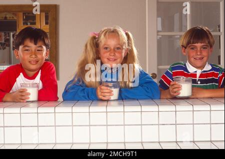 Trois enfants se tenant derrière un mur, chacun tenant un verre de lait. Tout en souriant à l'appareil photo Banque D'Images