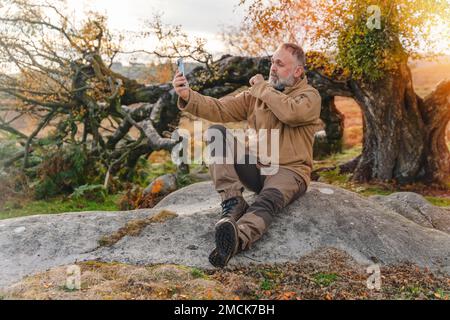 Homme barbu dans des bottes en forme de sphée atteignant la destination et se reposant sous l'arbre et prenant des photos au téléphone dans Peak District au coucher du soleil le jour d'automne Trav Banque D'Images