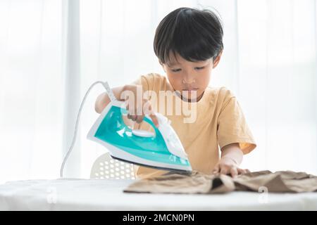 jeune garçon heureux fer son uniforme d'école avec fer sur planche à repasser à la maison pendant le week-end pour le nettoyage de la maison activité familiale Banque D'Images