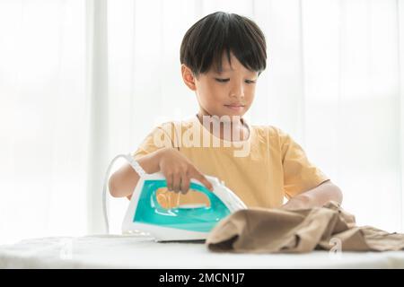 jeune garçon heureux fer son uniforme d'école avec fer sur planche à repasser à la maison pendant le week-end pour le nettoyage de la maison activité familiale Banque D'Images