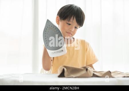 jeune garçon heureux fer son uniforme d'école avec fer sur planche à repasser à la maison pendant le week-end pour le nettoyage de la maison activité familiale Banque D'Images