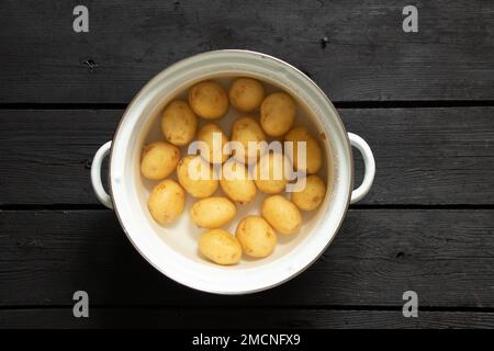 les jeunes pommes de terre brutes entières dans l'eau dans une casserole sont sur une table en bois noir, les pommes de terre dans une casserole, les faire bouillir Banque D'Images