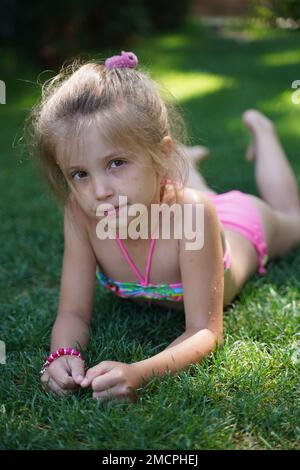 Une petite fille blonde dans un maillot de bain coloré se trouve sur l'herbe et regarde dans l'appareil photo gros plan. Vacances d'été. Mise au point sélective Banque D'Images