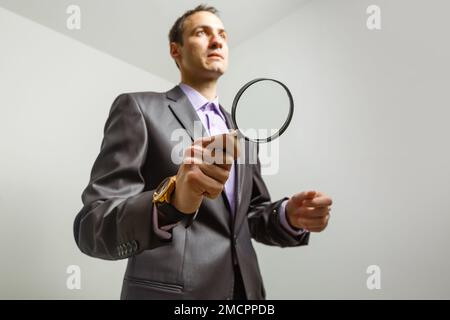 Homme d'affaires hippster sérieux en costume tenant la loupe et regardant la caméra, sur fond gris Banque D'Images