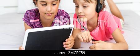 Deux petites filles mignonnes utilisent une tablette numérique et sourient lorsqu'elles sont allongées sur le lit dans la chambre des enfants Banque D'Images