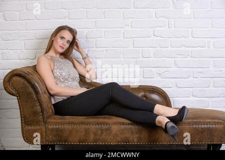 Jolie jeune femme avec cheveux longs, maquillage discret et une chemise pailletée sans manches, pantalon noir long et chaussures sur un canapé en cuir marron Banque D'Images