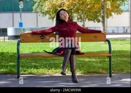 Jolie femme chinoise avec un pantalon collant noir et un manteau rouge est assise sur un banc de parc et profite du soleil en automne avec les yeux fermés et le regard rêveur Banque D'Images