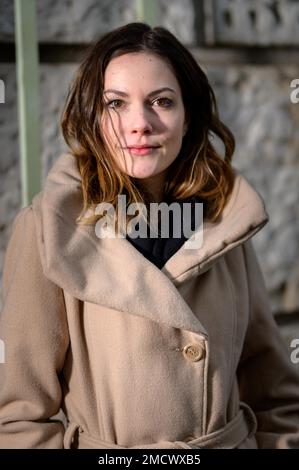 Jolie jeune femme avec de longs cheveux brunette, un manteau en tissu brun clair et une écharpe noire devant un mur de pierres naturelles Banque D'Images