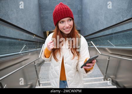 Portrait redhead fille touriste, monte les escaliers avec smartphone, suit l'itinéraire sur l'application de téléphone mobile, tient sac à dos et sourit Banque D'Images