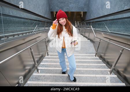 Portrait redhead fille touriste, monte les escaliers avec smartphone, suit l'itinéraire sur l'application de téléphone mobile, tient sac à dos et sourit Banque D'Images