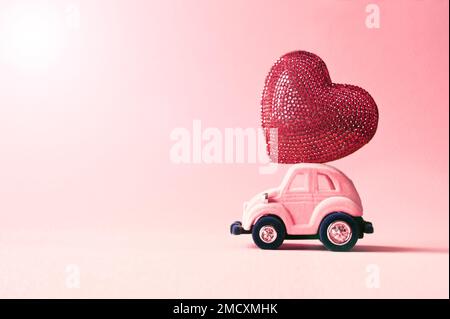 Voiture-jouet rétro offrant un coeur d'artisanat pour la Saint-Valentin sur fond rose Banque D'Images