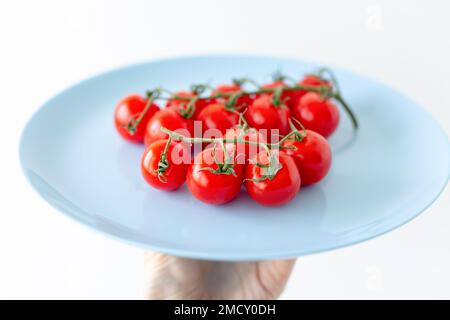 Des branches de tomates cocktail fraîches de cerise rouge vif se trouvent sur une assiette bleue sur un fond blanc sur la main d'un homme. Banque D'Images