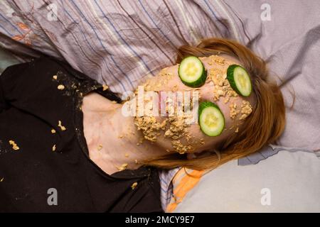 Une femme fait un masque cosmétique à la maison à partir de flocons d'avoine sur son visage et concombre.Traitements anti-âge, concept de soins de la peau. Banque D'Images