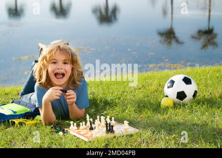 Gamin excité jouant au jeu d'échecs dans l'arrière-cour, en posant sur l'herbe. Échecs de jeu d'enfant concentré. Enfant jouant à un jeu de société en extérieur. Banque D'Images