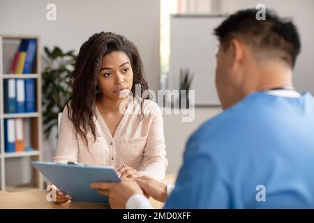 Un médecin coréen d'âge moyen en uniforme consulte une triste jeune femme afro-américaine montre un comprimé en clinique Banque D'Images