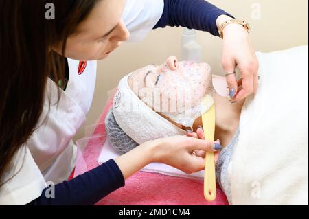 Un cosmétologue enlève un masque de feuille hydratant sur le visage d'une femme, procédures de cosmétologie. Banque D'Images