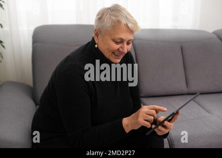 Une femme de race blanche souriante d'âge moyen s'assoit sur un canapé dans le salon et navigue sur Internet sans fil sur une tablette, une femme moderne et heureuse se détend sur un canapé à la maison Banque D'Images