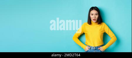 Portrait de la femme coréenne déçue dans un chandail jaune, frognant et regardant bouleversé, debout sur fond bleu Banque D'Images