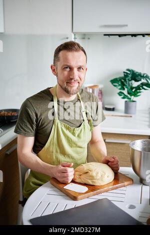 Beau boulanger barbu à la maison assis à la table posant. Caucasien beau portrait de boulanger mâle en tablier vert uniforme souriant et regardant l'appareil photo avec de la pâte sur bois Banque D'Images