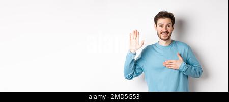 Homme souriant qui se soulève le bras et tient la main sur le coeur, faisant la promesse dire la vérité, jurer ou prêter serment, debout sur fond blanc Banque D'Images