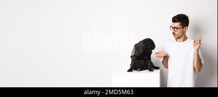 Beau jeune homme enseignant des commandes de chien, parlant à mignon pug noir, debout sur fond blanc Banque D'Images