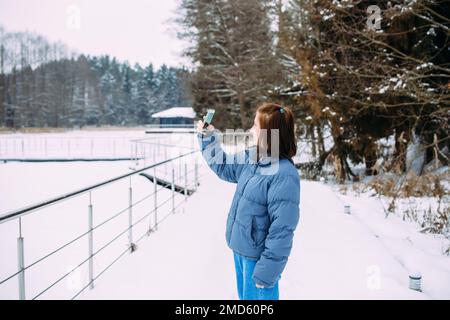 femme en hiver avec un téléphone prend un selfie dans la nature dans le parc par temps froid Banque D'Images