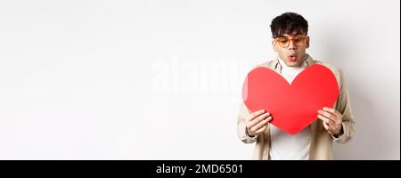 Un homme charmant et surpris reçoit une carte postale à cœur rouge le jour de la Saint-Valentin, en regardant le cadeau avec émerveillement, en appréciant le jour des amoureux, debout sur du blanc Banque D'Images