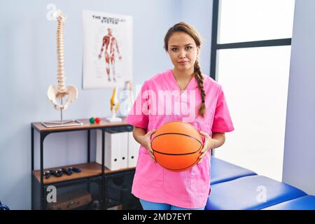 Jeune femme hispanique travaillant à la clinique de physiothérapie tenant le ballon de basket-ball détendu avec une expression sérieuse sur le visage. simple et naturel Banque D'Images