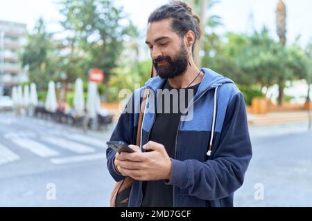 Jeune homme hispanique musicien utilisant un smartphone tenant un étui de guitare dans la rue Banque D'Images