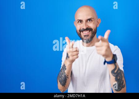 Homme hispanique avec tatouages debout sur fond bleu pointant les doigts vers l'appareil photo avec le visage heureux et drôle. bonne énergie et vibes. Banque D'Images