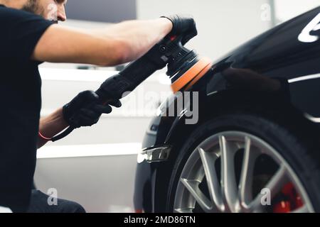 Vue à angle bas d'une voiture professionnelle détaillant un ouvrier à l'aide d'un polisseur orbital pour polir la peinture noire de la voiture. Roue et pneu dans le cliché. Photo de haute qualité Banque D'Images