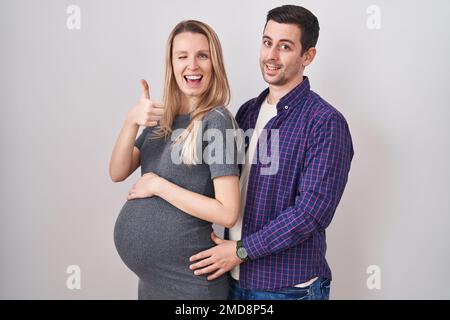 Jeune couple s'attendant à un bébé debout sur fond blanc faisant des pouces heureux vers le haut geste avec la main. approbation de l'expression en regardant la caméra showi Banque D'Images